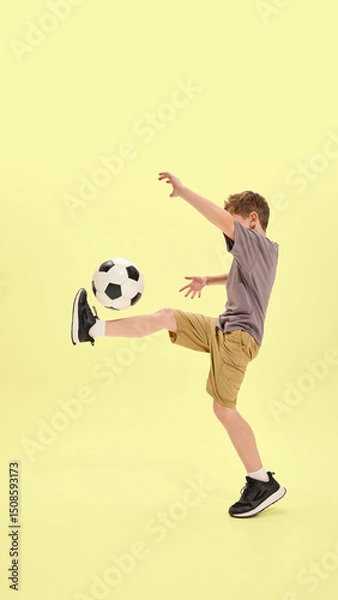 Fototapeta Boy lifting leg to balance ball in classic soccer move, wearing shorts and sneakers, yellow background enhances focus on movement and coordination. Concept of summer activities, hobby, holidays
