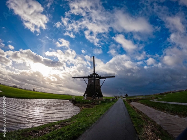 Fototapeta An ancient Dutch windmill used for pumping water on a blue sky with white clouds. Amazing landscape from the Netherlands at dusk in spring time 