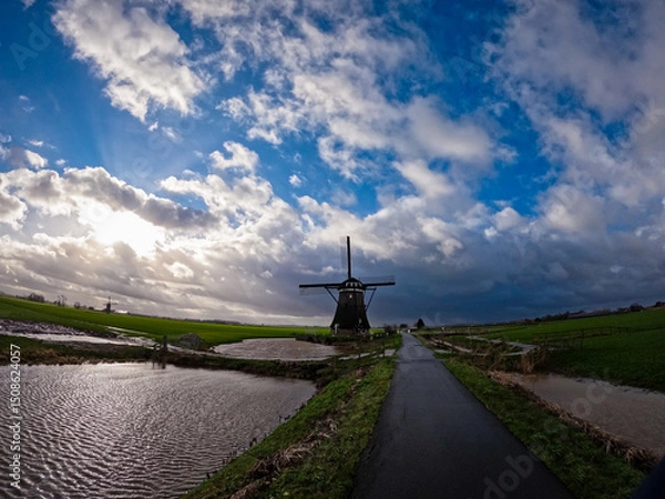 Fototapeta An ancient Dutch windmill used for pumping water on a blue sky with white clouds. Amazing landscape from the Netherlands at dusk in spring time 