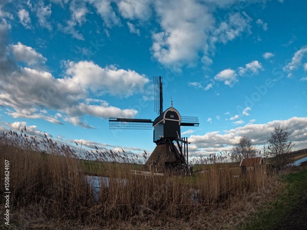 Fototapeta An ancient Dutch windmill used for pumping water on a blue sky with white clouds. Amazing landscape from the Netherlands at dusk in spring time 