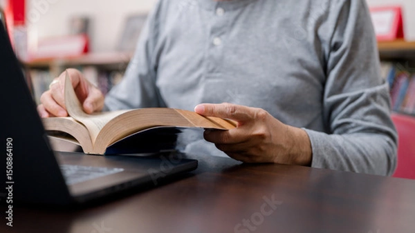 Fototapeta Close Up Person Focused On Book At Desk, Illuminated By Soft Lighting Near Library Open Book, Older Style, Laptop Create Studious Vibe Wearing Shirt, With Strong Focus In Study Setting
