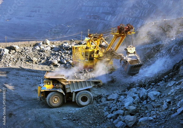 Obraz Excavator loading iron ore into heavy dump trucks on the opencast mining site