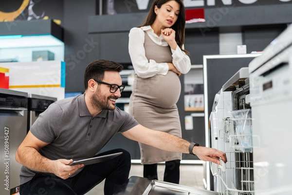 Obraz Smiling salesman holding a tablet, discussing and showing dishwashers to a pregnant customer, selecting the best options in a modern home appliance store