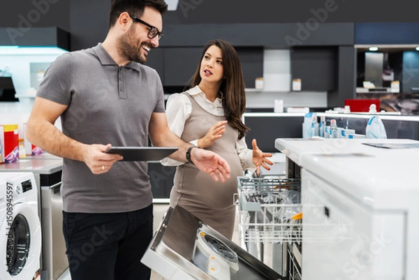 Obraz Smiling salesman holding a tablet, discussing and showing dishwashers to a pregnant customer, selecting the best options in a modern home appliance store