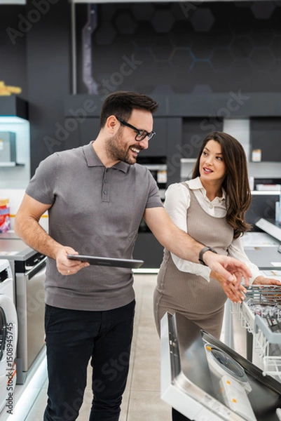 Obraz Smiling salesman holding a tablet, discussing and showing dishwashers to a pregnant customer, selecting the best options in a modern home appliance store