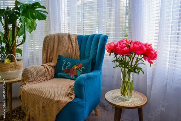 Fototapeta Bright pink peonies in a clear glass vase on a rustic wooden table, cozy interior with teal armchair, houseplants, and soft natural light. Homey and vibrant atmosphere.