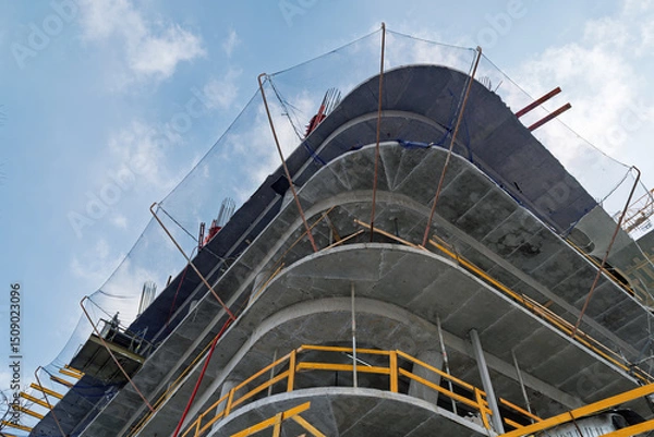Fototapeta Modern concrete building rises under a clear sky, safety nets and scaffolding showing progress and precision of urban construction.