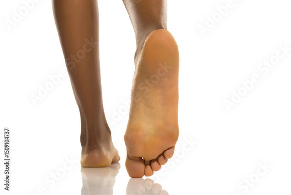 Fototapeta Close-up rear view of black woman's bare feet walking with visible sole, clean and healthy skin, studio shot on white background.