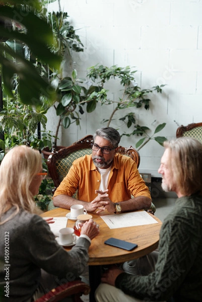 Fototapeta Group of three people sitting at round table, discussing plans in cozy indoor garden setting, surrounded by lush green plants, look relaxed and engaged in conversation