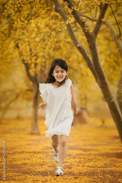 Obraz Happy little girl in white dress running in the park along  the blooming yellow trees, childhood memories and active kids concept