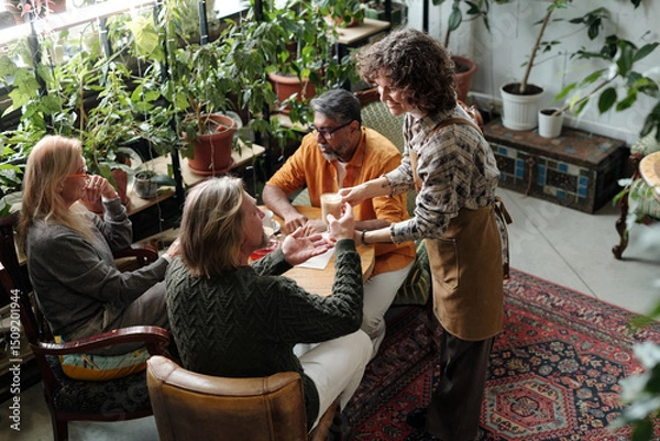 Fototapeta Group sitting around wooden table in cozy cafe surrounded by greenery, engaging in lively conversation and sharing tea while server hands small teacup to person wearing green sweater