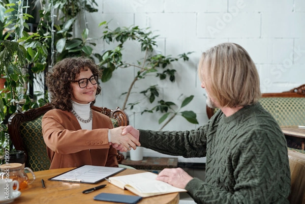 Fototapeta Two professionals shaking hands across a table in cozy, casual business setting with plants in background. Both have documents and notebooks on table, creating friendly and productive aura