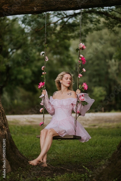 Fototapeta Woman in a Flowing Pink Dress Sitting on a Floral Swing in Nature