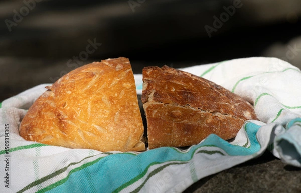 Fototapeta Close-up of two half and fresh loaves of bread lying on a kitchen towel. The bread is photographed outdoors.
