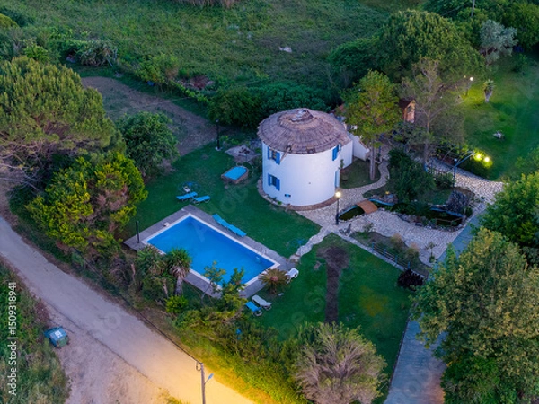 Fototapeta Aerial view of a quaint white windmill house with a swimming pool surrounded by lush greenery.