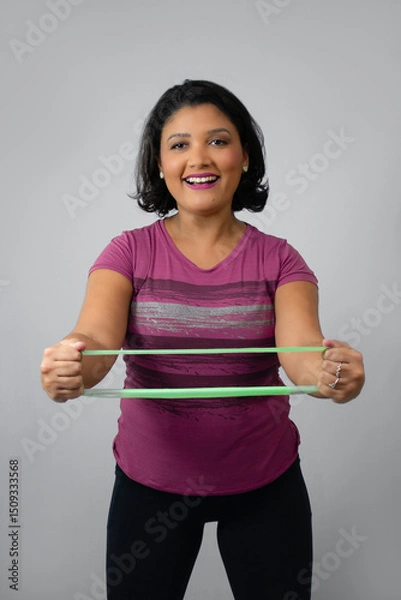 Fototapeta Happy fitness woman exercising arms with a green rubber band. Ideal for home workout, muscle strengthening and injury recovery. Studio portrait, neutral gray background with copy space.