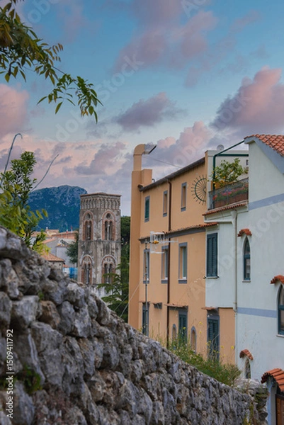 Obraz A stone bell tower with arched windows stands in Ravello, Italy, surrounded by pastel buildings, terracotta roofs, greenery, and a pink blue sky.
