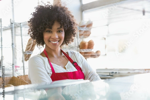 Obraz Baker wearing red apron smiling behind glass case in bakery, leaning by wire racks and windows