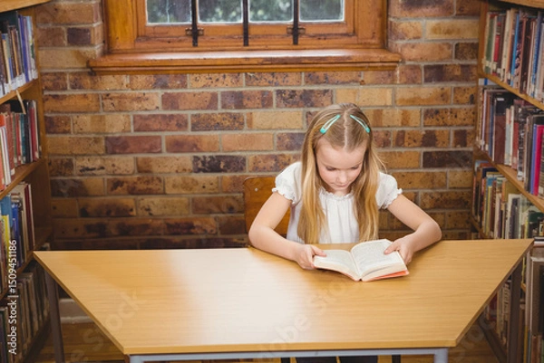 Fototapeta Girl reading paperback book at wooden desk in library with exposed brick wall and bookshelves