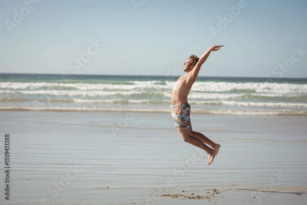 Fototapeta Man jumping above wet sand near shoreline at low tide, wearing colorful floral swim trunks