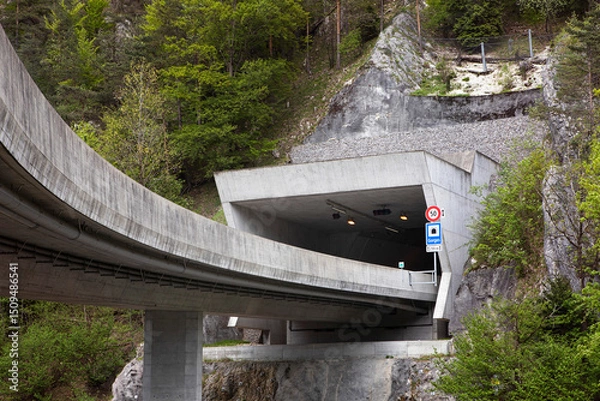 Fototapeta Entrance to a tunnel on a highway, pass trough mountain. Entry from flyover to road tunnel, highway in mountains, drive through mountain range and extreme terrain in Switzerland.