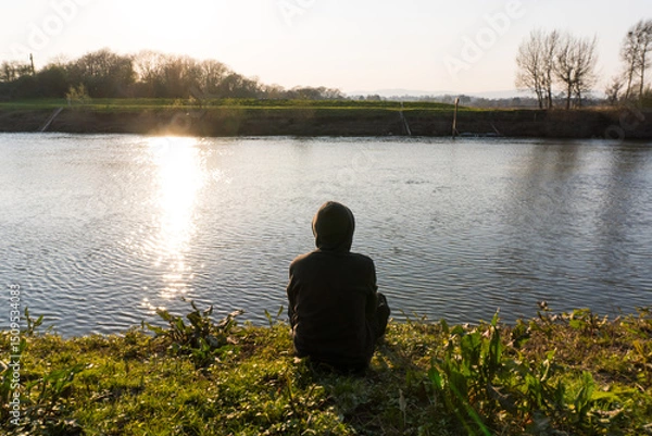 Fototapeta A lonely hooded, person, back to camera. Sitting by a river bank on a winters afternoon in the countryside