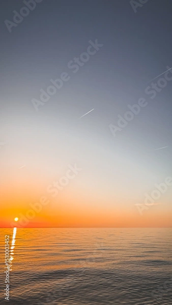 Fototapeta A serene sunset over calm ocean waters, with the sun low on the horizon and a gradient sky fading from orange to deep blue, vertical