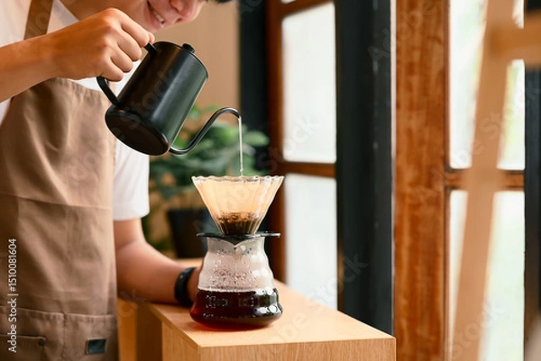 Fototapeta Young man in apron brewing coffee using the pour-over method in warm kitchen