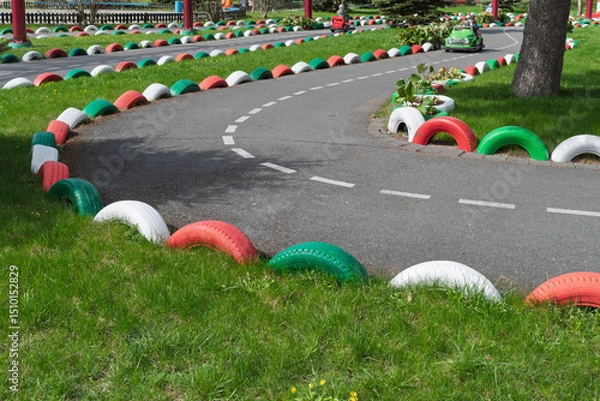 Fototapeta Ornamental racing track in park features winding asphalt road bordered by colorful tires on green grass, inviting playful movement.