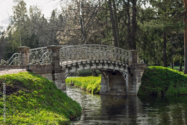 Fototapeta Elegant wrought iron bridge spans calm park canal, framed by greenery and old trees, offering serene atmosphere in spring sunlight.
