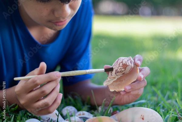 Fototapeta A young boy carefully cleans a seashell with a brush while lying on the grass, showcasing curiosity, nature appreciation, and a hands-on learning experienceideal for education or hobby concepts.