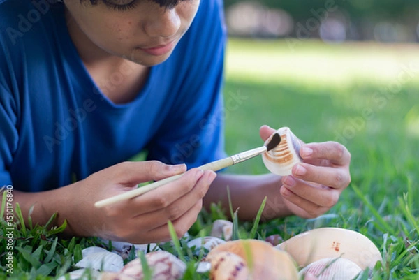 Fototapeta A young boy carefully cleans a seashell with a brush while lying on the grass, showcasing curiosity, nature appreciation, and a hands-on learning experienceideal for education or hobby concepts.