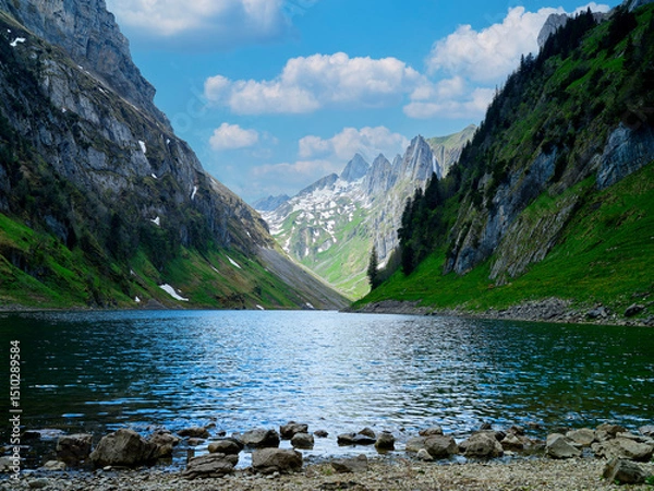 Fototapeta Scenic mountain lake surrounded by green hills and rocky cliffs under a blue sky with clouds