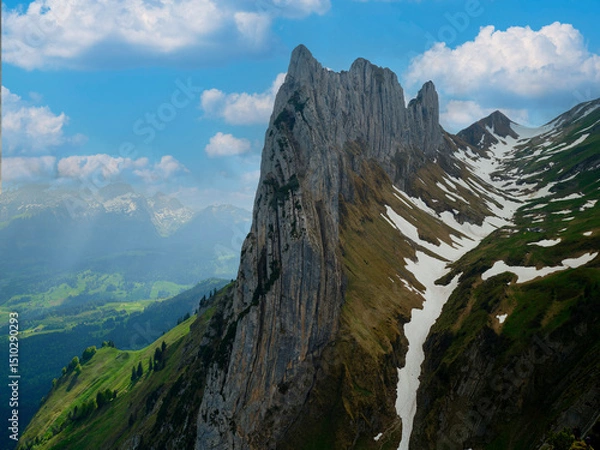 Fototapeta Majestic mountain peak rises above verdant hills and patches of snow under a bright blue sky filled with fluffy clouds