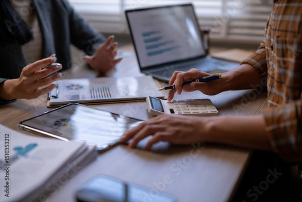Fototapeta Two people are sitting at a table with a laptop, a calculator, and a tablet