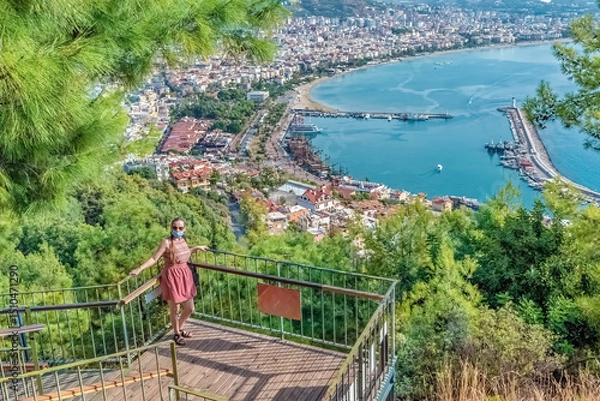 Fototapeta A woman stands on a viewpoint platform in Alanya, Turkey, overlooking the stunning coastline and harbor. The panoramic view showcases the Mediterranean Sea and surrounding mountains under a clear sky
