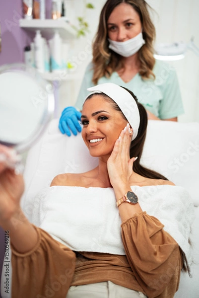 Obraz Beautiful and happy young woman sitting in medical chair and looking in the mirror. She is satisfied after successful beauty treatment with hyaluronic acid fillers or botulinum toxin injections.