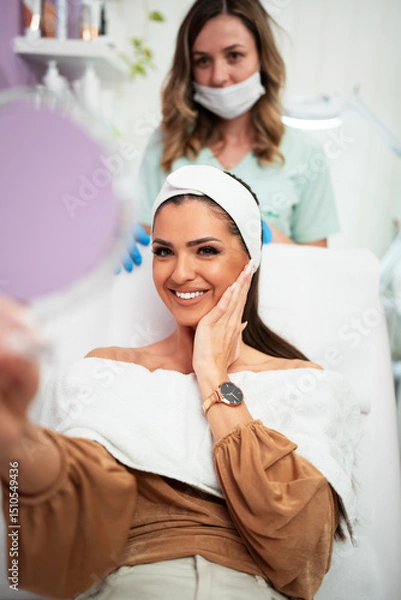 Obraz Beautiful and happy young woman sitting in medical chair and looking in the mirror. She is satisfied after successful beauty treatment with hyaluronic acid fillers or botulinum toxin injections.