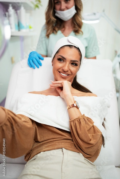 Obraz Beautiful and happy young woman sitting in medical chair and looking in the mirror. She is satisfied after successful beauty treatment with hyaluronic acid fillers or botulinum toxin injections.
