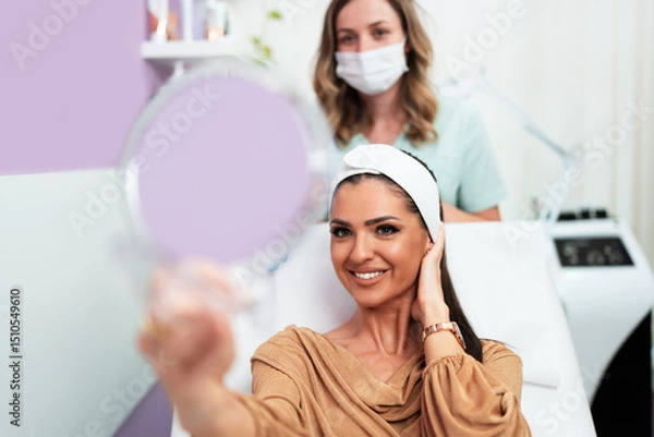 Obraz Beautiful and happy young woman sitting in medical chair and looking in the mirror. She is satisfied after successful beauty treatment with hyaluronic acid fillers or botulinum toxin injections.