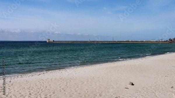 Fototapeta Scenic view of a quiet sandy beach and calm sea waves under a clear blue sky. A breakwater structure with buildings and a watchtower stretches along the horizon in the distance.
