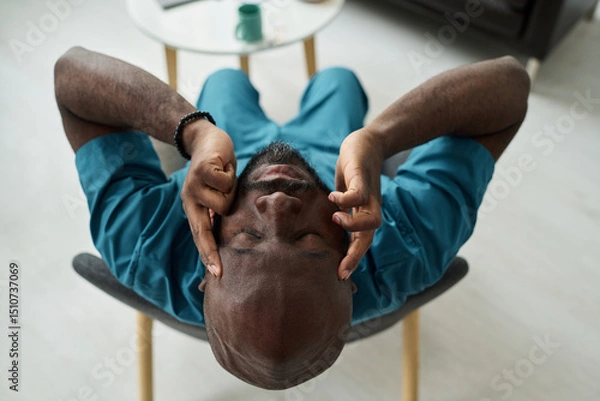 Fototapeta Exhausted medical professional in uniform rubbing eyes while sitting in chair with table in background, indicating stress and fatigue from work in healthcare environment