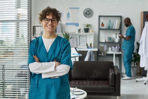 Fototapeta Healthcare worker standing with crossed arms in a medical office, smiling. Background includes a coworker working on documents and medical equipment, creating a professional environment