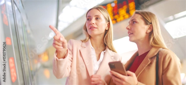Fototapeta Two women at a train station comparing the travel data between smartphone and info display