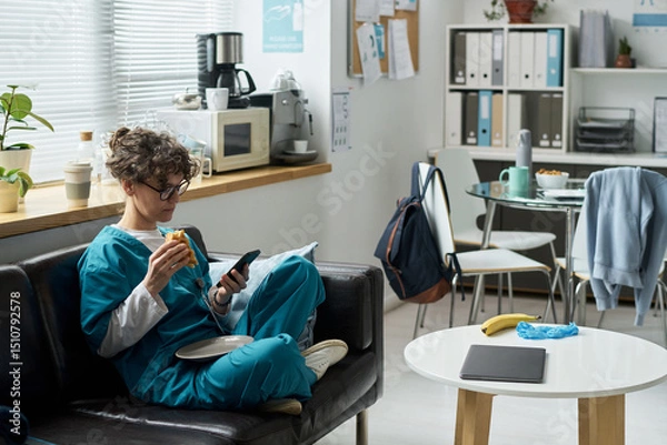 Fototapeta Nurse relaxing in break room, holding phone and snack. Tables filled with food and drinks, office supplies organized on shelves. Natural light filtering through window enhancing ambiance