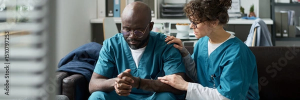 Fototapeta Nurse comforting a distressed healthcare worker sitting in a break room. Medical uniforms and professional workspace suggesting a healthcare setting
