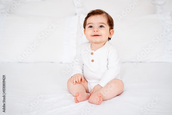 Fototapeta a baby on a white bed at home, a place for text, a cute smiling little child getting ready for bed, a portrait of a little boy child