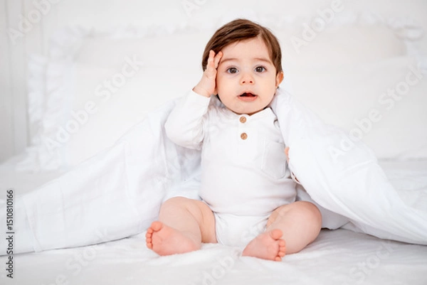 Fototapeta A baby under a blanket on a white bed at home, a place for text, a cute smiling little child getting ready for bed