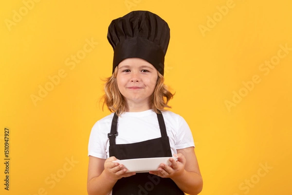 Fototapeta Funny child cook with cooking plate. Chef kid boy making healthy food. Portrait of little child in chef hat isolated on studio background. Child chef. Cooking process.