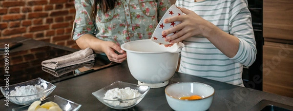 Fototapeta Unrecognizable young daughter pours flour into mixing bowl while baking with his mother in modern kitchen, surrounded by ingredients, sharing warm moment of culinary learning and family connection.
